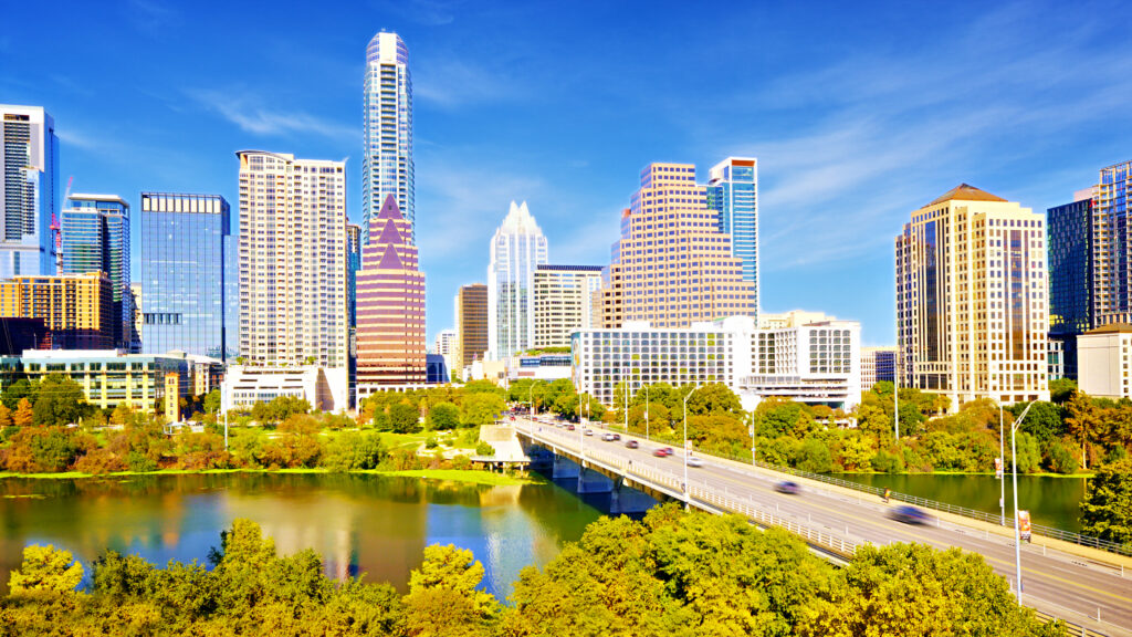 Daytime photo of Austin skyline, with a bridge over Ladybird Lake in the foreground.
